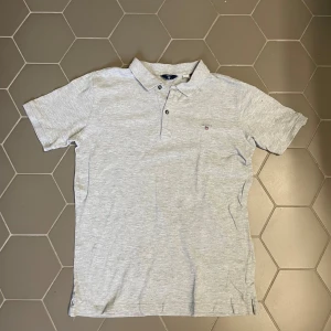 Ljusgrå pikétröja från GANT - Säljer en stilren ljusgrå pikétröja från GANT med klassisk krage och två knappar framtill. Tröjan har korta ärmar och en liten broderad GANT-logga på bröstet. Perfekt för en clean och avslappnad look.