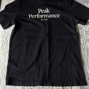 Svart Peak Performance t-shirt - Svart t-shirt från Peak Performance med vit logga och text på bröstet. Klassisk passform med korta ärmar och rund hals. Tillverkad i mjuk bomull som känns skön mot huden. Perfekt för en clean och stilren look.