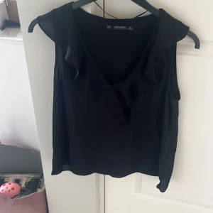 Svart ärmlös blus från Zara Basic - Säljer en svart ärmlös blus från Zara Basic Collection. Toppen har v-ringning med volangdetalj framtill och är tillverkad i ett lätt och luftigt polyestertyg. Perfekt för dig som gillar stilrena och enkla plagg med en liten twist.