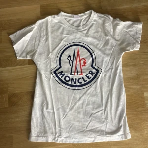 Vit Moncler t-shirt med stor logga - Vit t-shirt från Moncler med stor, ikonisk logga i blått och rött framtill. Klassisk rund halsringning och korta ärmar. Tillverkad i mjuk bomull för en skön känsla. Perfekt för dig som gillar stilrena och trendiga plagg.