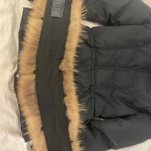 Svart pufferjacka med päls från Hollies - Säljer en svart pufferjacka från Hollies med avtagbar stor brun päls. Jackan har flera fickor med dragkedja och knappar, samt snygga detaljer som markerad midja och orange insida. Perfekt för kalla dagar när du vill vara både varm och stilren. Tyvärr är detaljen på dragkedjan lite förstörd då materialet är gjort av tråd. jackan är köpt för 5 tusen förra vintern men säljs just för 3000kr priset kan diskuteras. 