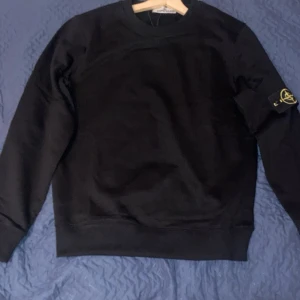 Svart sweatshirt från Stone Island - Svart sweatshirt från Stone Island med klassisk rund hals och ikonisk logotyp-patch på ärmen. Tröjan har långa ärmar och ribbade muddar vid ärmslut och nederkant. Tillverkad i mjuk bomull för en skön och stilren look. Passar xs och s