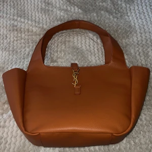 Brun läder handväska från Yves Saint Laurent - Snygg brun handväska från Saint Laurent i mjukt skinn med guldigt YSL-spänne framtill. Väskan har breda handtag och en praktisk innerficka med dragkedja. Perfekt för dig som vill ha en stilren och lyxig accessoar med ikonisk logga.