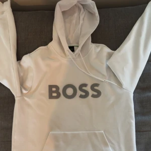 Vit hoodie från BOSS med logga - Säljer en stilren vit hoodie från BOSS med stor grå logga framtill. Tröjan har huva med dragsko och en klassisk känguruficka. Perfekt för dig som gillar en clean och modern look. Materialet är mjukt och skönt, troligtvis bomullsmix.