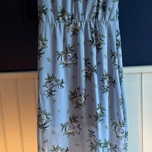 Ljusblå blommig långklänning - Superfin ljusblå långklänning med blommigt mönster i grönt och blått. Klänningen har resår i midjan för en bekväm passform och är ärmlös, perfekt för varma dagar. Tunt och luftigt material som känns skönt mot huden.