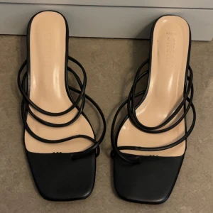 Svarta sandaler med remmar från Shegan - Stilrena svarta sandaler från Shegan med flera tunna remmar och fyrkantig tå. Snygg minimalistisk design med öppen tå och låg klack, perfekta för sommaren. Tillverkade i syntetmaterial med beige innersula.