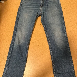 Blå jeans från Tiger of Sweden - Snygga blå jeans från Tiger of Sweden med klassisk femficksdesign och diskreta slitningar framtill och baktill. Jeansen har raka ben och normal passform, tillverkade i mjukt denimtyg med orangea sömmar och läderpatch bak i midjan.