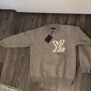 Grå stickad Louis Vuitton tröja  - Grå stickad tröja från Louis Vuitton med stort vitt LV-logo både fram och bak. Tröjan har rund halsringning, ribbade muddar och är långärmad. Storleken är M men passar både L och tröjan är helt ny.