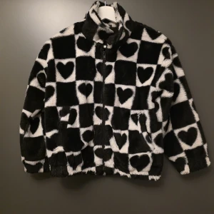 Svartvit teddyjacka med hjärtan - Fluffig teddyjacka i svart och vitt med rutigt mönster och hjärtan. Jackan har hög krage, dragkedja framtill och långa ärmar. Perfekt för dig som vill sticka ut med en mysig och trendig look.