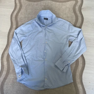 Ljusblå skjorta från Tiger of Sweden - Snabb leverans.                                                              Säljer en stilren ljusblå skjorta från Tiger of Sweden, modell Brodie. Skjortan har extra slim passform, klassisk krage och knäppning framtill. Tillverkad i bomull och passar perfekt till dig som gillar en clean och snygg look.