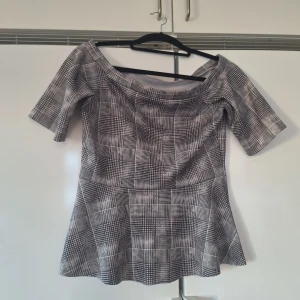Rutig svartvit blus med vid hals stl M - Snygg blus i svartvitt rutigt mönster med bred, vid halsringning och korta ärmar. Modellen har en lätt utställd nederdel som ger en feminin siluett. Perfekt för dig som gillar grafiska mönster och stilren look.