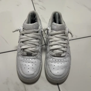 Nike Air Force 1 vita sneakers - Klassiska Nike Air Force 1 sneakers i helvitt skinn med lågt skaft och rund tå. Skorna har perforerade detaljer på tån, breda vita snören och den ikoniska Air-sulan. Perfekta för dig som gillar en clean och tidlös streetstil.