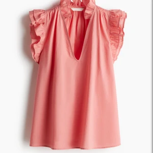 Rosa blus med volangärmar - Supersöt rosa blus med djup v-ringning och volangdetaljer vid ärmar och hals. Blusen är ärmlös och har en luftig passform, perfekt för varma dagar. Tillverkad i mjuk bomull som känns skön mot huden.