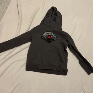 Mörkgrå hoodie från Zavetti Canada - Säljer en mörkgrå hoodie från Zavetti Canada med stor broderad logga och röd lönnlöv på bröstet. Tröjan har huva med tryckt text, känguruficka och långa ärmar. Perfekt för dig som gillar streetwear och snygga detaljer.