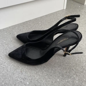 Svarta pumps från Dolce & Gabbana - Säljer ett par eleganta svarta pumps från Dolce & Gabbana med spetsig tå och smal klack. Skorna har en snygg rem med guldfärgat spänne. Perfekta för dig som gillar klassisk och stilren design.