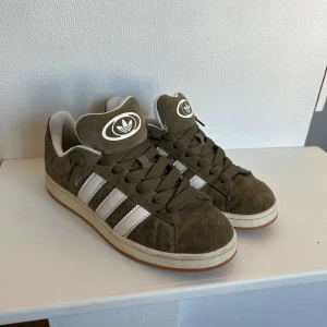 Adidas Campus olivgrön  - Snygga Adidas campus i olivgrön mocka med klassiska vita ränder på sidorna och vit sula. Skorna har rund tå, snörning och gummisula. Perfekta för dig som gillar retrostil och vill ha något som sticker ut lite extra.