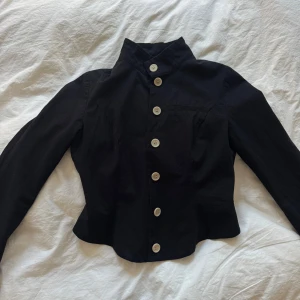 Svart kavaj från Baum und Pferdgarten - Unik svart kavaj med hög krage och vita knappar framtill från Baum und Pferdgarten. Jackan har lång ärm, figurnära passform och en cool detalj i ryggen med blå- och vitrandigt tyg. Tillverkad i bomull och fodrad invändigt.