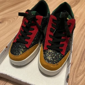 Harry Potter glitter sneakers Hogwarts - Färgglada sneakers med Hogwarts-emblem och Harry Potter-tryck på sulan. Skorna har svart glitter, röda och senapsgula mockadetaljer, blå och gröna partier samt svarta skosnören. Perfekta för dig som älskar magi och vill sticka ut med en unik stil.