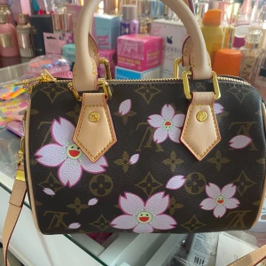 Louis Vuitton x Murakami blommig handväska - Superfin handväska från Louis Vuitton x Murakami 20th Anniversary med ikoniskt monogram och rosa-blommigt mönster. Väskan har ljusa handtag, guldfärgade detaljer och dragkedja. Insidan är fodrad i rosa tyg och har exklusiv märkning.