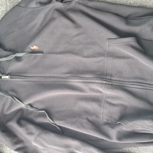 Svart hoodiejacka från Polo Ralph Lauren - Svart hoodiejacka från Polo Ralph Lauren med dragkedja framtill, huva med snörning och klassisk röd broderad logga på bröstet. Jackan har stora fickor framtill och är tillverkad i mjuk bomull för en skön och avslappnad stil.