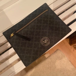 Svart kuvertväska från By Malene Birger - Snygg svart kuvertväska från By Malene Birger med diskret rutigt mönster och guldfärgad dragkedja. Väskan har en stilren design med logga framtill och är gjord i tåligt syntetmaterial. Perfekt att bära till alla tillfällen när du vill ha med det viktigaste.