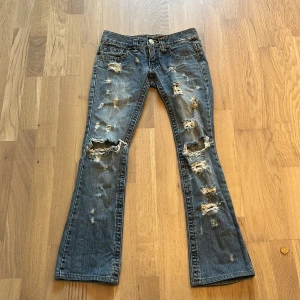 Unika blå bootcut jeans från TMX  - Low waist Bootcut jeans från TMX Jeans Cloth med slitna och trasiga detaljer. Jeansen har klassisk femficksdesign, låg midja och är tillverkade i denim. Perfekt för dig som gillar en edgy och avslappnad stil. Äkta från 2000 talet