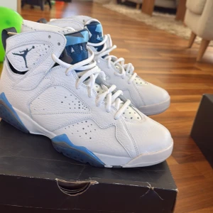Nike Air Jordan 7 Retro UNC vit/blå - Nike Air Jordan 7 Retro UNC i vitt och blått skinn med snygga detaljer som Jumpman-logga på sidan och siffran 23 på hälen. Skorna har högt skaft, snörning och perforerade partier för extra stil. Perfekta för dig som vill sticka ut med klassiska basketvibbar.