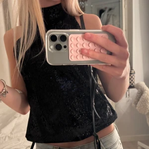 Svart paljettopp med hög hals - Säljer en svart ärmlös topp med hög hals och glittriga paljetter över hela tyget. Toppen har en rak passform och är perfekt för dig som vill sticka ut. Snygg att styla med jeans eller kjol för en cool look.