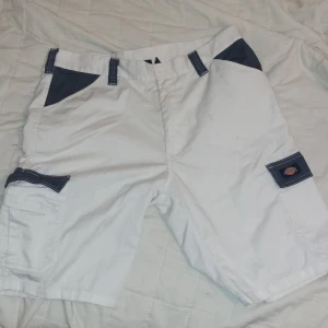 Vita Dickies cargo shorts med blå detaljer - Snygga vita cargo shorts från Dickies med kontrasterande mörkblå detaljer på fickor och hällor. Klassisk loose passform, flera praktiska fickor och tydlig Dickies-logga på benet och bakfickan. Perfekta för en chill sommarstil. Har ett litet hål som är sytt framtill, som jag fick från skateboarding, du kan se det på sista bilden