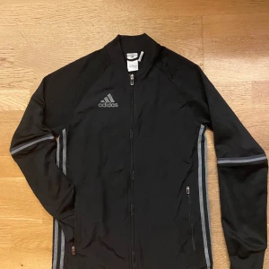 Svart Adidas vindjacka med grå detaljer - Svart vindjacka från Adidas med grå ränder längs ärmarna och logga på bröstet. Jackan har hel dragkedja framtill, två sidofickor med dragkedja och långärmad design. Perfekt för sportiga dagar och streetwear.