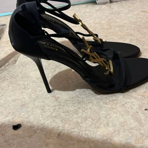 Svarta Saint Laurent klackar med YSL-detalj - Snygga svarta klackar från Saint Laurent med tunna remmar och ikonisk YSL-logga i guld framtill. Skorna har öppen tå, hög smal klack och är tillverkade i skinn. Perfekta för dig som vill sticka ut med en lyxig och elegant look. Alla storlekar finns😍höjd: 8cm och 10cm