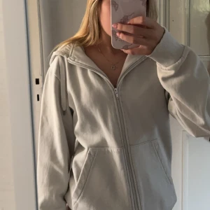 vit hoodie med dragkedja - Säljer en vit hoodie med dragkedja och huva. Tröjan har långa ärmar, ribbade muddar och två stora fickor framtill. Perfekt för en chill och avslappnad stil.