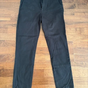 Svarta kostymbyxor Riley slim fit - Svarta kostymbyxor från Riley i slim fit-modell. Byxorna har snedställda fickor fram och två bakfickor med knapp. Materialet är en mix av ull, bomull, modal och elastan för en skön och stretchig känsla.