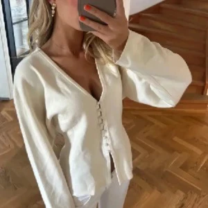 Vit kofta med knappar och v-ringning - Säljer en stilren vit kofta med v-ringning och dekorativa knappar framtill. Koftan har långa ärmar och en något figurnära passform, perfekt att styla till både jeans och kjol. Materialet känns mjukt och bekvämt.