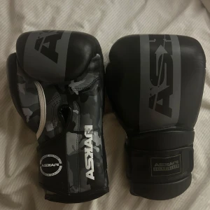 Svarta boxningshandskar från Askari - Säljer ett par svarta boxningshandskar från Askari med grått camouflagemönster och tydlig logga. Handskarna är tillverkade i skinn och har bred kardborrestängning runt handleden för extra stöd. Perfekta för dig som vill ta din träning till nästa nivå.