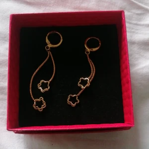 Guldiga örhängen med blommor - Säljer ett par snygga guldiga örhängen med hängande kedjor och små blommor i öppen design. De har en modern och trendig look, perfekta för att lyfta din outfit. Kommer i en röd, quiltad ask.