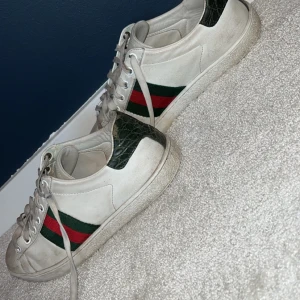 Gucci Ace vita sneakers med gröna detaljer - Gucci Ace sneakers i vitt skinn med klassiska gröna och röda ränder på sidorna. Baksidan har gröna detaljer med reptilmönster. Snygg vågig sula med unikt mönster undertill. Perfekta för dig som vill ha en ikonisk och trendig look.