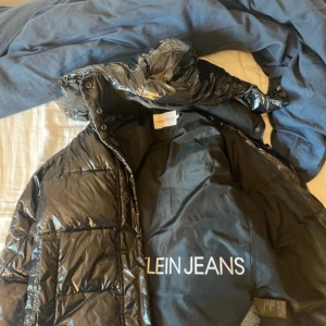Svart pufferjacka Calvin Klein Jeans M - Snygg svart pufferjacka från Calvin Klein Jeans med glansig finish och huva. Jackan har CK-logga på bröstet, dragkedja framtill och quiltad design. Perfekt för kalla dagar när du vill hålla stilen och värmen.