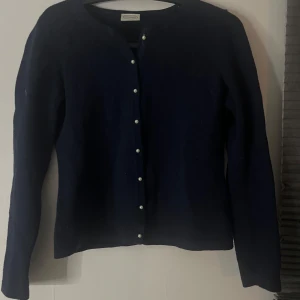 Mörkblå kofta från Newhouse med pärlknappar - Snygg mörkblå kofta från Newhouse med långa ärmar och pärlknappar framtill. Klassisk och stilren modell i mjukt material, perfekt att slänga över en t-shirt eller klänning. Enkel rund halsringning och normal passform.