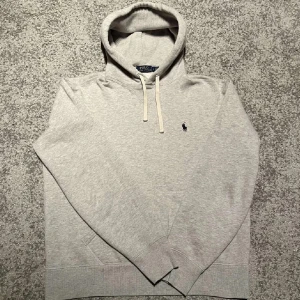 Grå hoodie från Polo Ralph Lauren - Snygg ljusgrå hoodie från Polo Ralph Lauren med klassisk broderad logga på bröstet. Tröjan har huva med snörning, känguruficka och är tillverkad i mjuk bomullsmix. Perfekt för chill dagar eller när du vill ha en clean och stilren look.