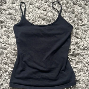 Svart enkel linnetopp med smala band - Svart basic linnetopp med smala axelband och rundad halsringning. Toppen är figurnära och tillverkad i mjuk bomull, perfekt att styla lager på lager eller bära som den är. Enkel och stilren design utan mönster eller detaljer.