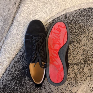Svarta sneakers från Christian Louboutin - Säljer ett par svarta sneakers från Christian Louboutin i mocka med klassisk röd sula och snörning. Skorna har rund tå och platt sula, samt diskreta sömdetaljer som ger en clean och lyxig vibe. Perfekta för dig som vill sticka ut med exklusiva detaljer. Utan box