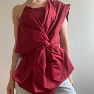 Vinröd asymmetrisk topp med knyt - Säljer en vinröd, asymmetrisk topp med stor knutdetalj framtill och en axel bar. Toppen har dragkedja i ryggen och är tillverkad i ett slätt tyg. Perfekt för dig som vill sticka ut med en unik och trendig look