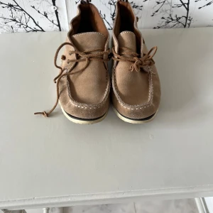 Beige mocka seglarskor Peak Performance - Snygga beige seglarskor från Peak Performance i mocka med vita kontrastsömmar och bruna snören. Skorna har en platt sula och randigt innerfoder för extra stilpoäng. Perfekta för dig som gillar en avslappnad men trendig look.