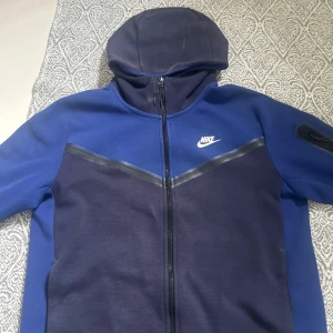 Blå Nike tech - Nike hoodie i två nyanser av blått med hel dragkedja och huva. Tröjan har lång ärm, vit Nike-logga på bröstet och en snygg V-detalj framtill. Perfekt för chill eller träning, med mjukt material och sportig look.