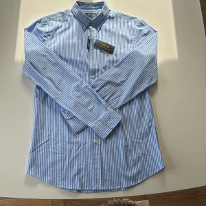 Polo Ralph Lauren Blåvit randig skjorta  - Snygg blå och vit randig skjorta från Polo Ralph Lauren i slim fit. Skjortan har klassisk button down-krage, långa ärmar och knappar framtill. Tillverkad i stretchig bomull för extra komfort. Perfekt för dig som gillar stilren och fräsch look.