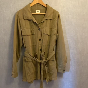 Beige overshirt med bälte från Indiska - Stilren grön tunn jacka från Indiska med knappar framtill, fyra stora fickor och knytskärp i midjan. Jackan har klassisk krage och långa ärmar med knapp vid ärmslut. Perfekt för lager-på-lager och chill stil.