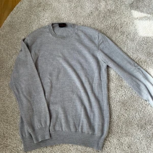 Grå merinoullströja från Oscar Jacobson - Säljer en stilren grå tröja från Oscar Jacobson i storlek M. Tröjan har en klassisk design med rund hals och långa ärmar. Perfekt för en avslappnad men ändå elegant look.