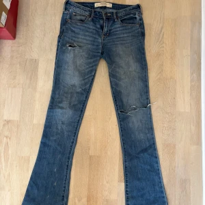 Blå bootcut jeans Abercrombie & Fitch - Snygga blå jeans från Abercrombie & Fitch med bootcut passform. Jeansen har slitna detaljer och hål på benen för en cool, avslappnad look. Klassiska bakfickor med dekorativ söm och läderpatch med logga bak i midjan. Storlek 26/31!🩷