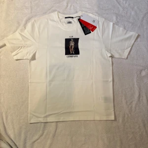 Vit C.P. Company t-shirt med print - Vit t-shirt från C.P. Company med tryck av en figur och logga på bröstet samt stort grafiskt print och texten 'April, 2024' på ryggen. Klassisk passform, rund hals och mjukt bomullsmaterial. Helt ny med tag kvar. Perfekt för dig som gillar streetwear med unik design. Storleken är M och passar bra på modellen som är 179 65kg. Skickar eller möts upp i Sthlm 